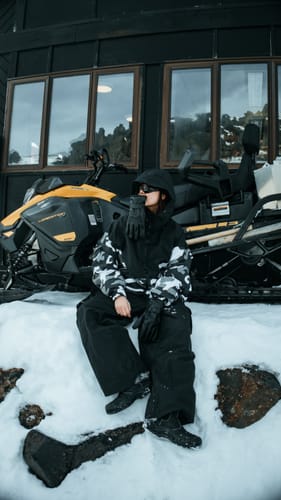 Customer's Women’s Black Baggy Cargo Snow Pants worn by a person sitting in snow near a snowmobile.