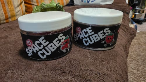 Kundenfoto von zwei THC Space Cubes Dosen mit weißem Deckel und schwarzem Etikett, platziert auf einer braunen Couch.