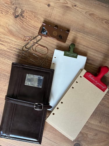 Customer's brown Infinite Quiet journal, loose pages, and paper clips laid out on a wooden surface.