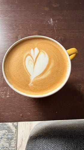 Customer's India Monsooned Malabar coffee in a yellow mug with heart-shaped latte art foam on a wooden table.