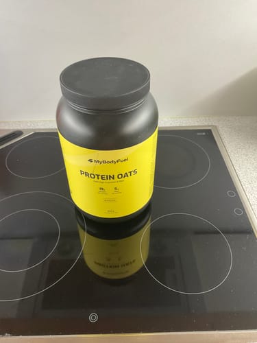 Kundenfoto der MyBodyFuel Protein Oats Dose in Schwarz und Gelb, stehend auf einem schwarzen Herd.