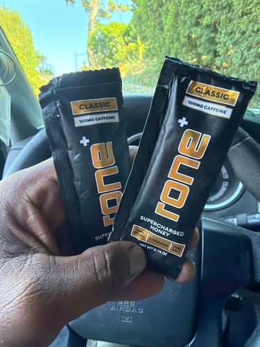 Customer's hand holding two black, rectangular SuperCharged Honey Caffeine packets inside a car.