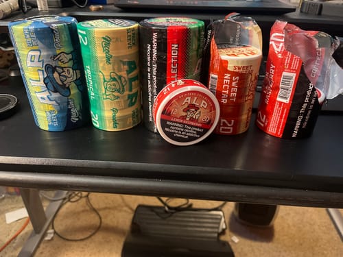 Reviewer's collection of Drifters nicotine pouches, including a centered Lemon Raspberry tin on a black surface.