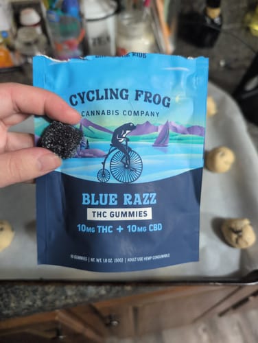 Reviewer's hand holding a dark-colored Blue Razz gummy in front of the matching product pouch.