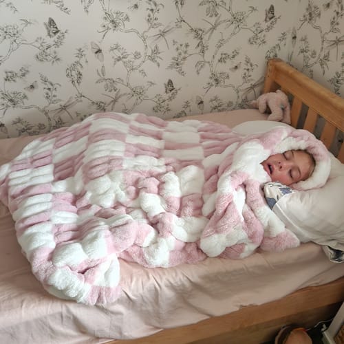 Customer's cozy Hugs Check Pink Blanket wrapped around a sleeping person on a bed with patterned wallpaper.