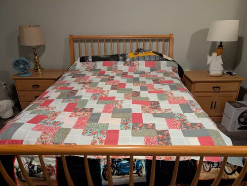 Reviewer's finished Mixmaster Manchester quilt draped over a bed with pink, blue, and green patterns.