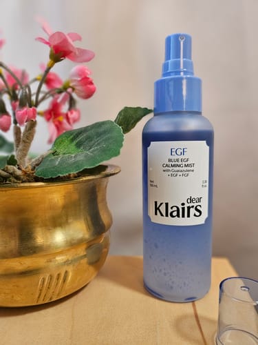 Customer's Blue EGF Calming Mist bottle standing on a wooden surface next to a decorative golden pot with pink flowers.