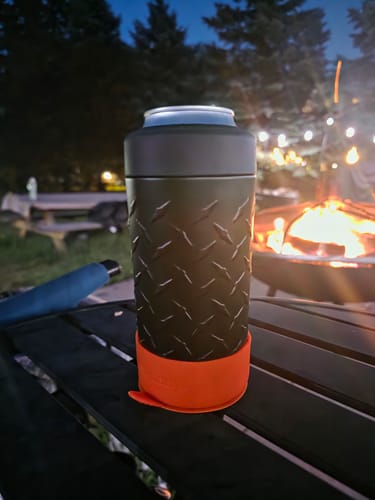 Customer's black Diamond Plate Universal Buddy can cooler with orange silicone base on a table by a fire pit.