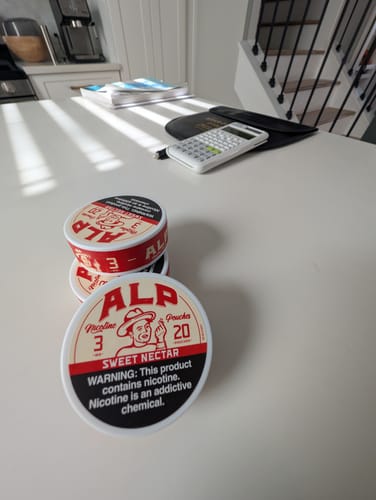 Customer's stack of round Sweet Nectar Nicotine Pouches tins on a table near a calculator.
