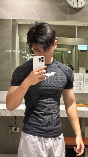 Reviewer wearing a fitted black Batman compression tee with armored pattern, taken as a mirror selfie in a gym.