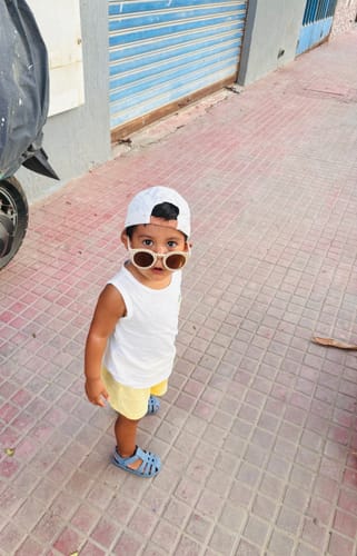 Customer's child wearing beige Nenina & Co sunglasses, a white cap, and a white tank top outdoors.