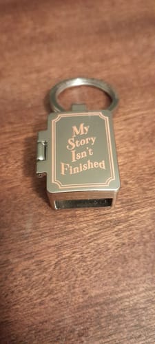 Customer's My Story Isn't Finished Zoxlox keychain, a metallic, book-shaped charm on a wooden surface.