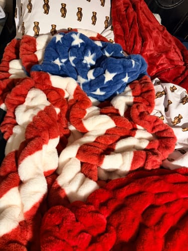 Customer's Hugs Flag Blanket draped on a bed with a bear print pillow and red plush blanket nearby.