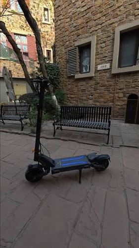 Customer's Ausom L2 Max Dual Motor E-scooter parked on a stone patio in front of a stone building with metal benches.