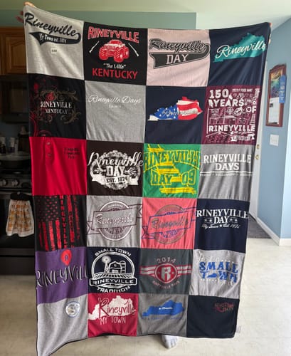 Customer's Project Repat T-Shirt Quilt held up, featuring various colorful patches showcasing Rineyville community themes.