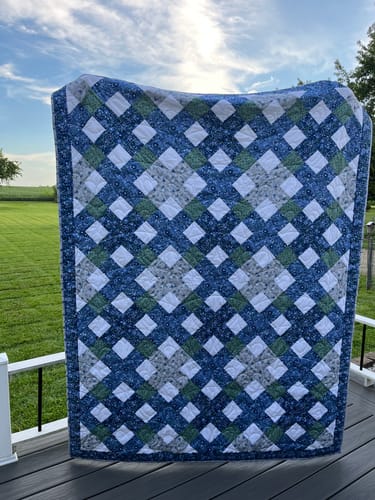 Customer's completed Belvoir Blue Precut Quilt Kit with a lattice design in blue, white, gray, and green hung outdoors.
