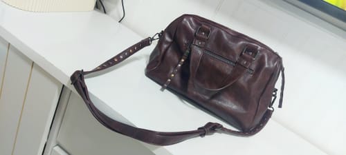 Customer's Cartera De Hombro Tachas Street en color café, mostrando las tachas en la correa sobre una superficie blanca.
