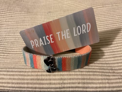 Customer's Hearts Charm attached to a colorful striped bracelet resting on a card that reads Praise the Lord