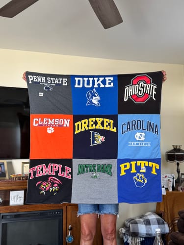 Customer's Project Repat T-Shirt Quilt featuring a grid of various college logos, held up indoors.
