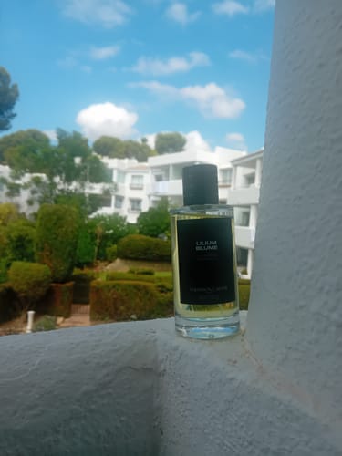 Customer's Lilium Blume perfume bottle on a balcony railing with a sunny garden and buildings in the background.