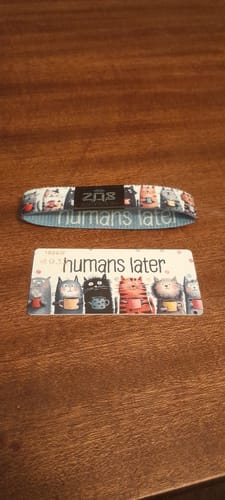 Reviewer's Humans Later bracelet and matching art card with cat illustrations on a wooden surface.