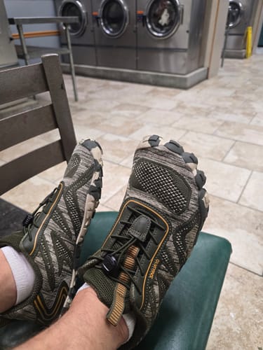 Reviewer wearing comfortable Active II Pro barefoot shoes with a camouflage pattern in a laundromat.