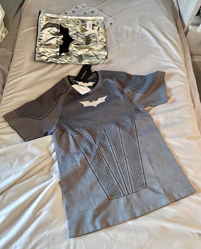 Customer's gray Batman Compression Tee with armored pattern on a bed, with metallic packaging in the background.