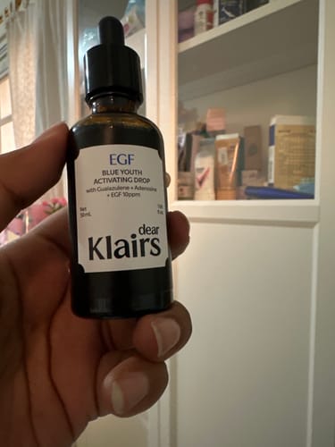 Reviewer's hand holding a bottle of Dear Klairs EGF Blue Youth Activating Drop serum against a blurry background.