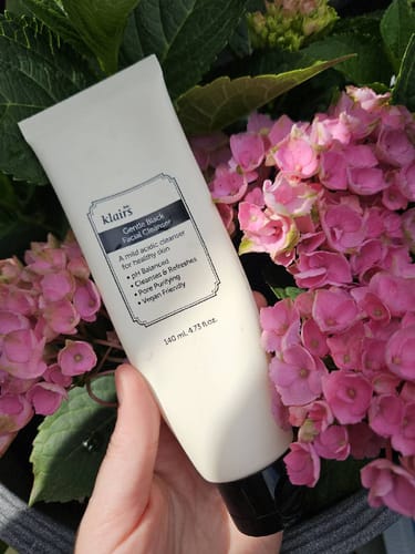 Customer's Gentle Black Facial Cleanser in a hand, held in front of bright pink hydrangea flowers.