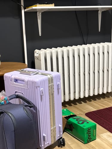Customer's light purple All-in-One Carry-On suitcase standing on a wood floor next to a dark bag and a soda box.