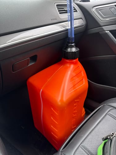 Customer's bright orange jug with a blue spout nozzle, placed on the front passenger seat of a car.
