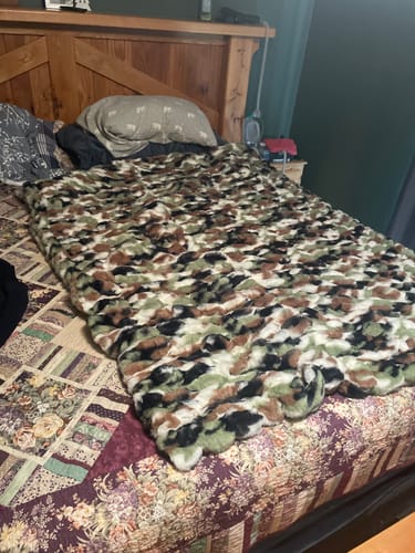 Customer's Hugs Pine blanket with camo-patterned plush texture spread over a bed with a wooden headboard.