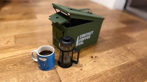 Customer's Coffee Resupply open olive green ammo tin next to a french press and coffee mug on a wooden floor.