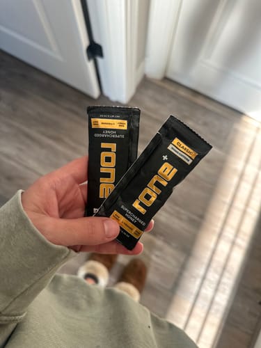 Customer's hand holding two slim black Rone Supercharged Honey caffeine packets.