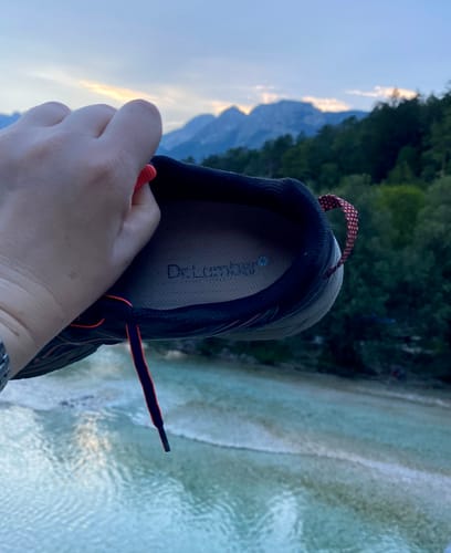Reviewer's hand holding a dark-colored trail shoe with an Ortopéd Pro Talpbetét inserted, outdoors by a river.
