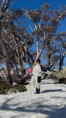Customer's off-white waterproof snow pants worn while posing with a snowboard in a snowy, sunlit forest.