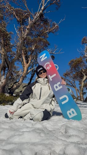 Customer's off white waterproof snow jacket worn by a person sitting in the snow holding a colorful snowboard.
