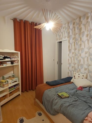 Customer's Mini Néo Applique en bois éclairée, suspendue dans une chambre avec des rideaux orange et un lit.