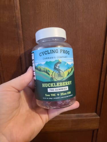 Customer's hand holding a jar of Huckleberry Gummies with a blue label featuring a frog on a penny-farthing bicycle.