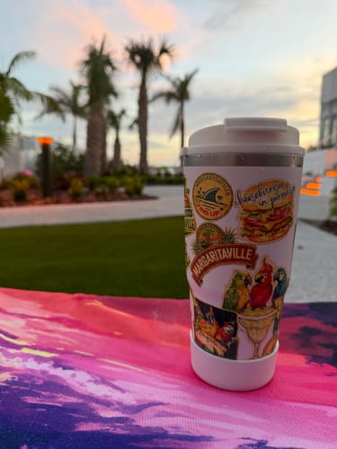 Reviewer's Margaritaville To-Go Buddy tumbler featuring decorative stickers in a tropical setting at sunset