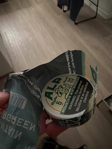 Customer's hand holding a round container of Mountain Wintergreen nicotine pouches with a partially torn wrapper.