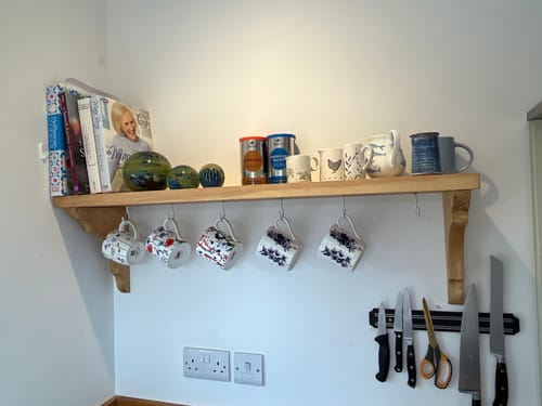 Customer's solid wood shelf mounted on a kitchen wall, holding books, mugs, and decor with hanging mugs below.