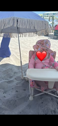 Customer's Amy portable booster chair V2 with a pink patterned bucket hat, sitting on a beach under a fringe umbrella.