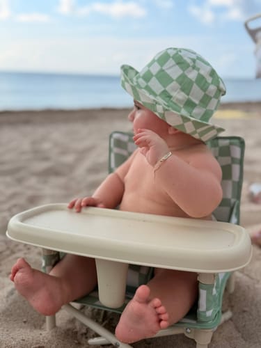 Customer's baby sitting in a Parker portable booster chair on a sandy beach, wearing a green checkerboard sun hat.