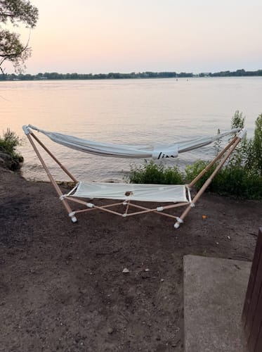 Customer's The Go Hammock on a metal frame set up on a sandy shoreline overlooking a lake at sunset.
