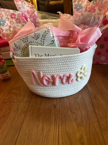 Customer's white woven basket customized with pink lettering reading Nora and a flower icon, filled with gifts.