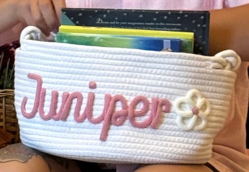 Customer's personalized Baby Knits knitted basket in white with pink name Juniper and a flower icon, filled with books.