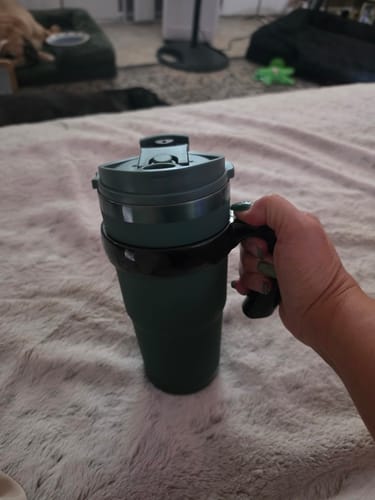 Customer's green To-Go Buddy cup held by a hand, showing its handle and lid on a soft textured surface.