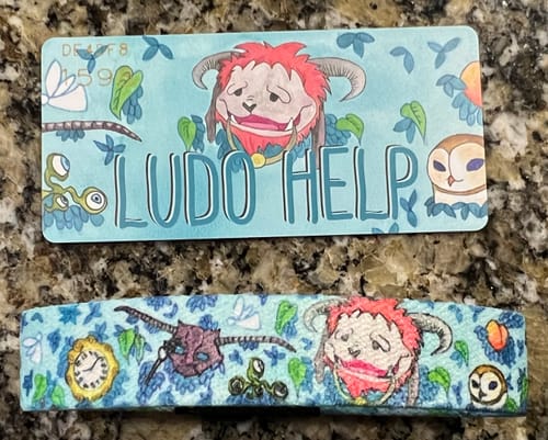 Reviewer's Ludo Help Labyrinth Bracelet next to its printed display card on a stone surface.