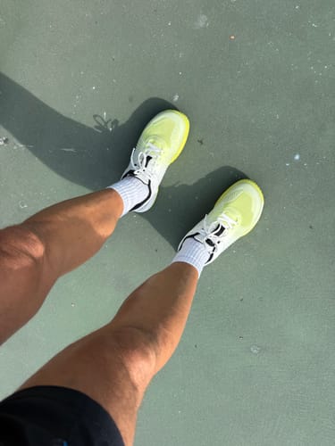 Reviewer's feet wearing neon yellow and white CUURT Momentum court shoes, standing on a tennis court.